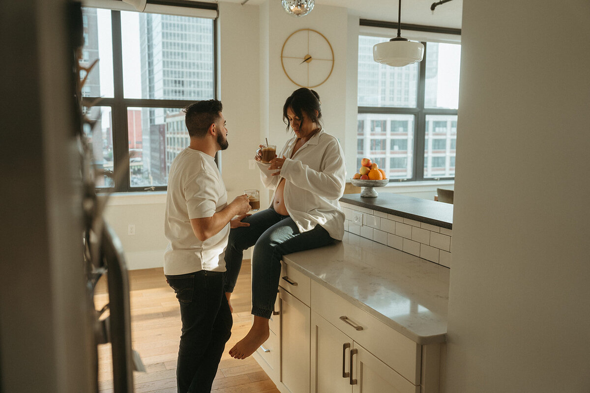 pregnant woman sits on kitchen counter with husband standing in front during maternity photos captured by NYC maternity photographer Elsie Goodman 