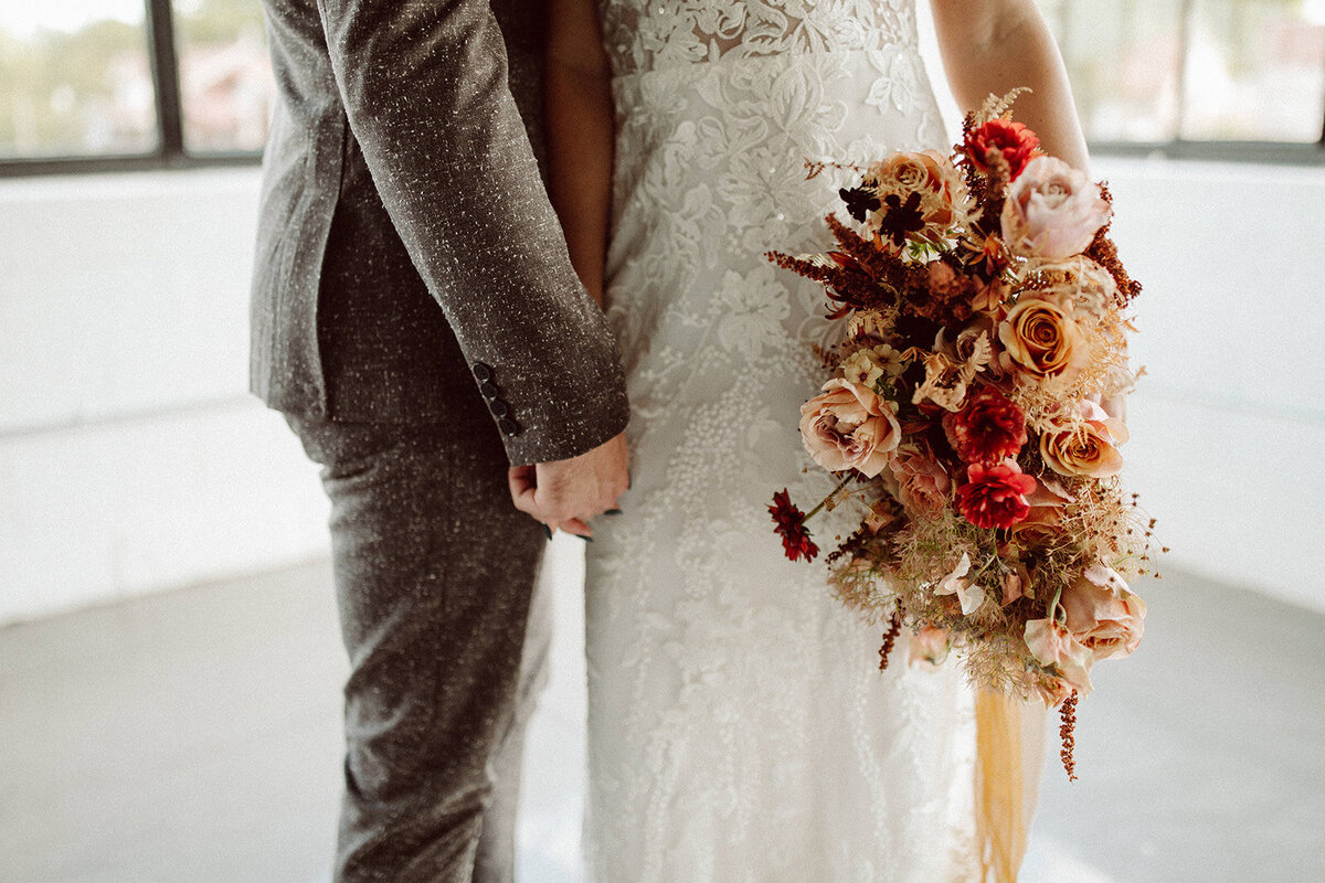 Close up of a bride and groom wearing a gray suit and white wedding gold holding hands and a bouquet of blush, cream and wine-colored flowers.