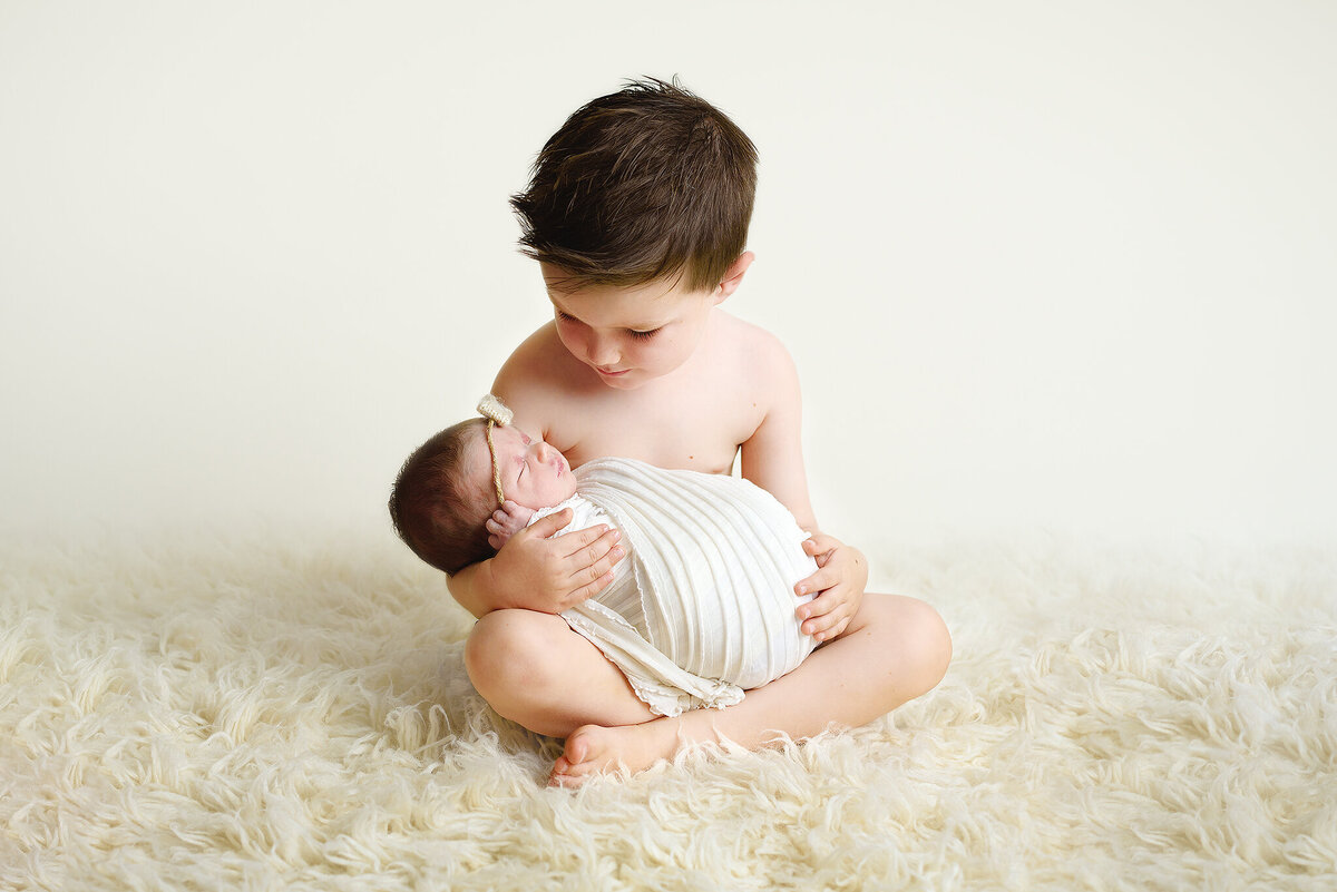 Big brother holding swaddled baby sister in lap with cream backdrop and rug in Jacksonville, FL.