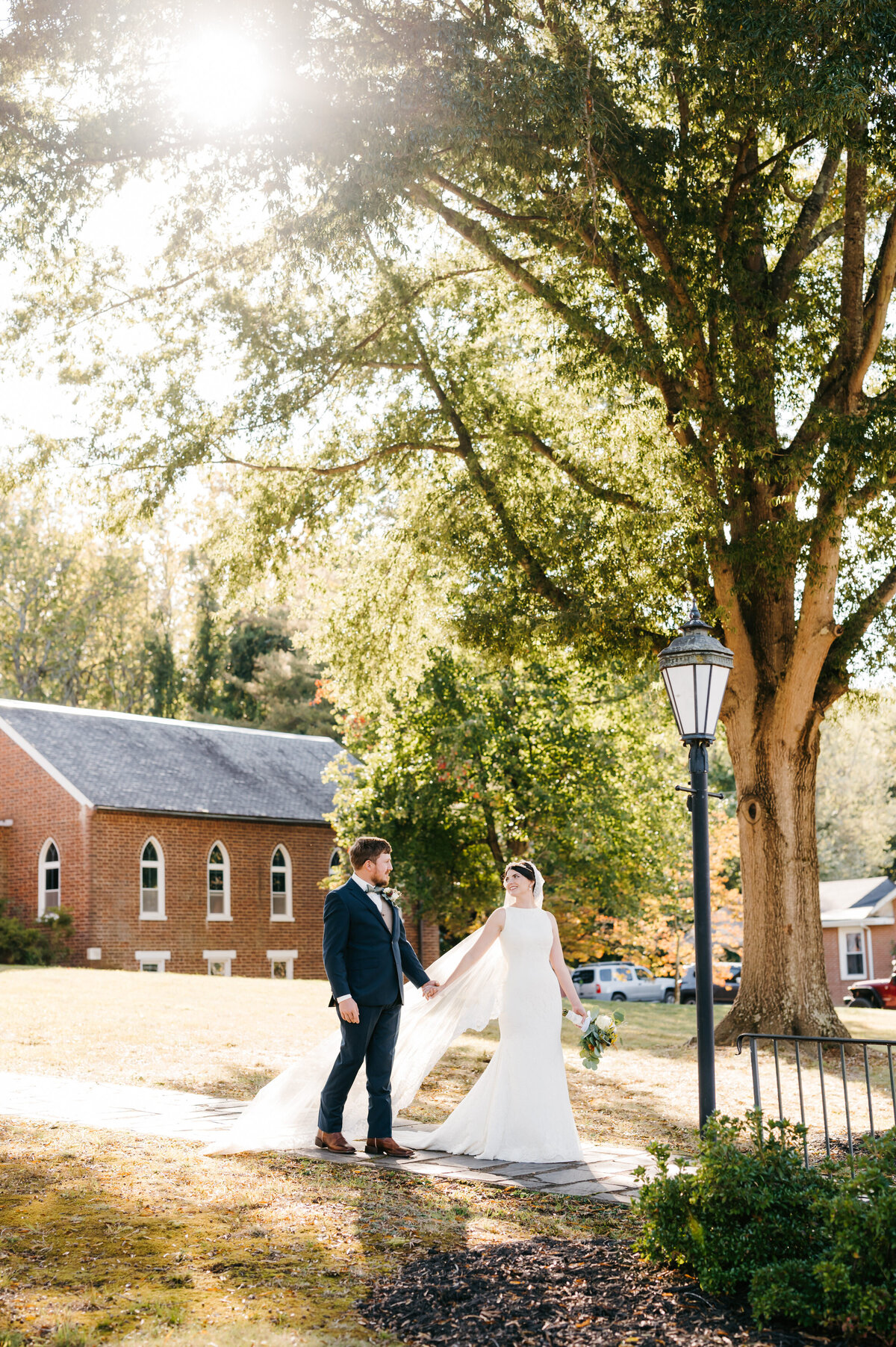 Richmond wedding photographer captures bride and groom exiting their chapel wedding while holding hands and smiling to one another as the sun shines down through the trees