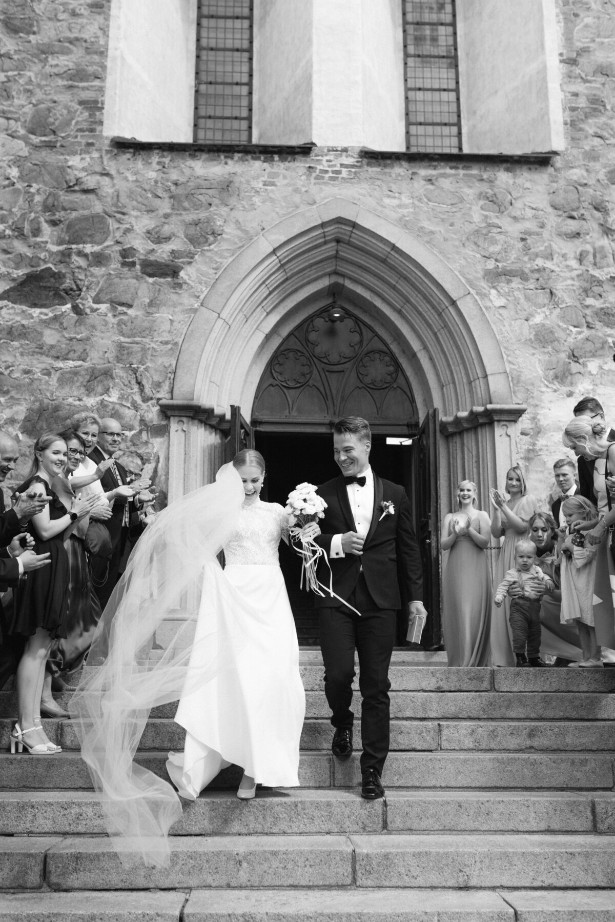 Wedding couple walking down the stairs of Turku cathedral after ceremony. Guests are cheering. Documentary wedding photography by photographer Hannika Gabrielsson.