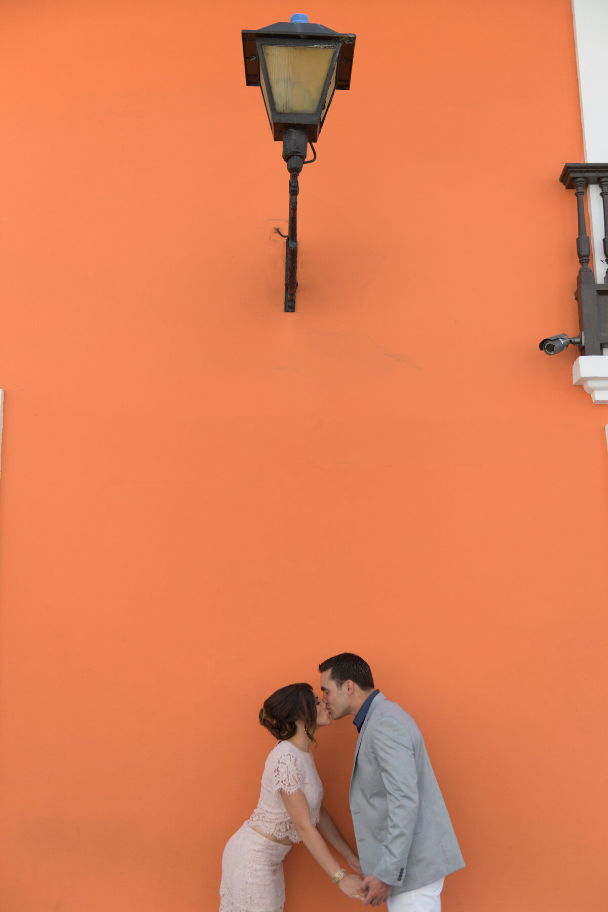 Playful engagement portrait of a couple kissing by a vibrant orange wall, captured by Rodrigo Varela Photography in Miami, Florida. Modern and artistic shot showcasing love and color.