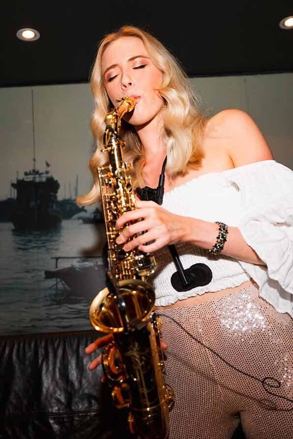 Blonde female saxophonist playing live to guests at an indoor event wearing a white off-shoulder top and glistening sequin pants.