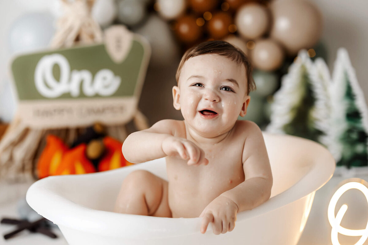 A smiling baby sits in a white bathtub amidst a camping-themed backdrop, captured by a talented portrait photographer from Bloomington, Illinois. Trees and a campfire decoration set the scene, accompanied by a charming sign that reads "One." Perfect for family photography enthusiasts!.