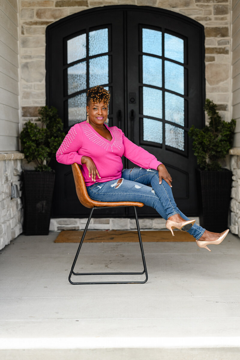 A black woman in a bright pink  sweater is sitting on a stool in front of a set of black doors during a branding photo shoot by a branding photographer in Denver.
