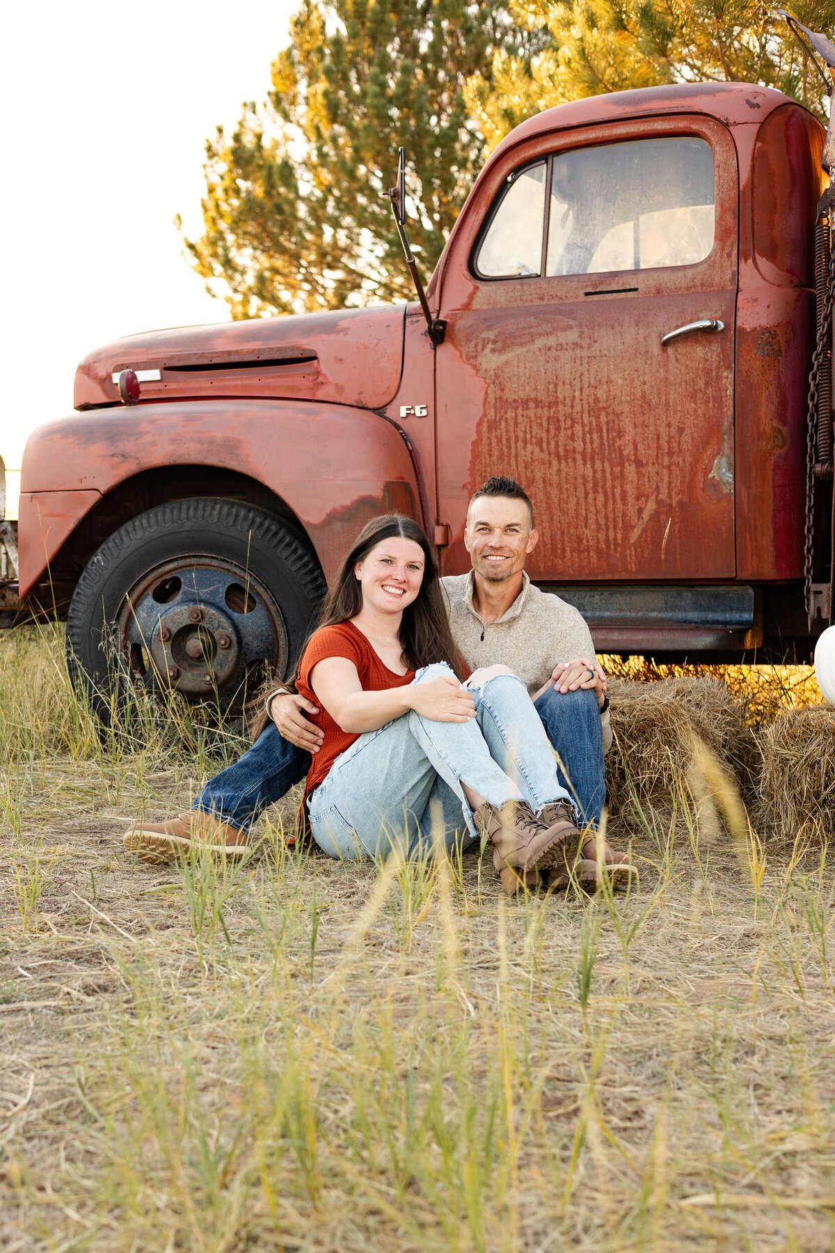 A husband and wife sit in the grass in front of a vintage red farm truck and smile at the camera.