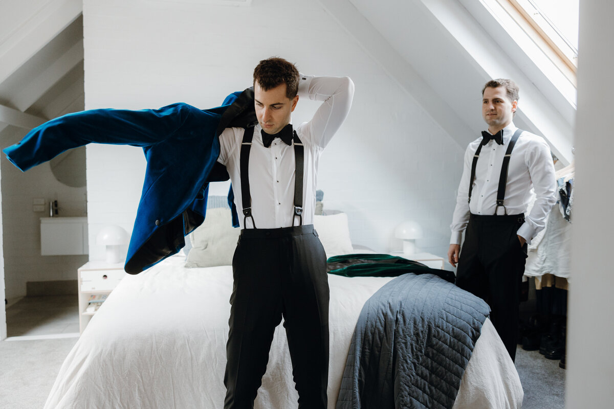 A groom wearing his blue velvet suit as his partner watches him.