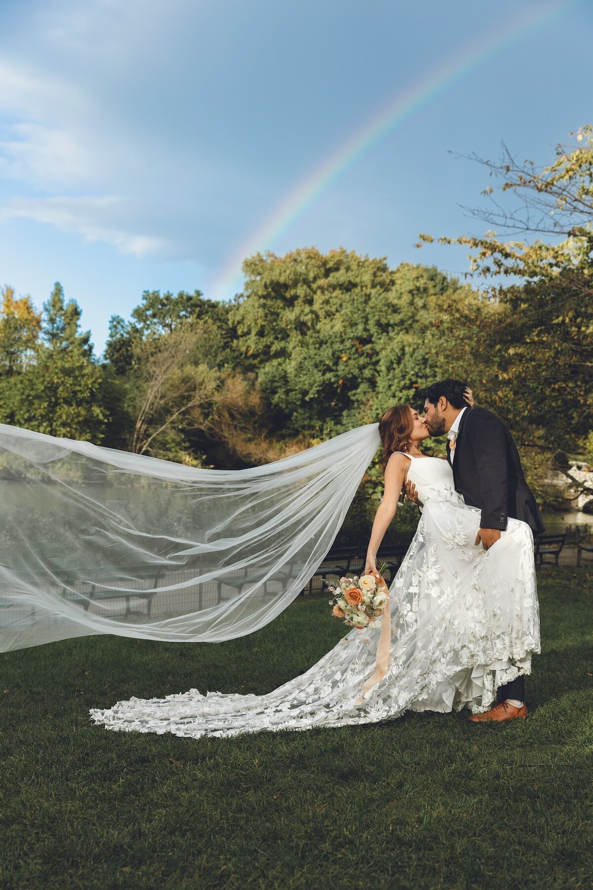 Groom kissing bride as veil floats in Central Park rainbow backdrop