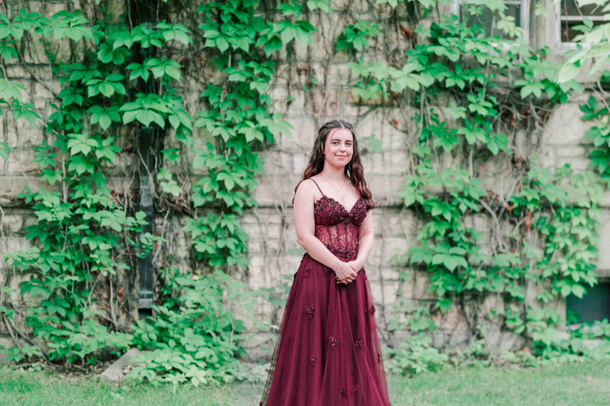 A beautiful young girl standing in a burgundy dress standing in front of the ivy wall on campus in Saskatoon.  