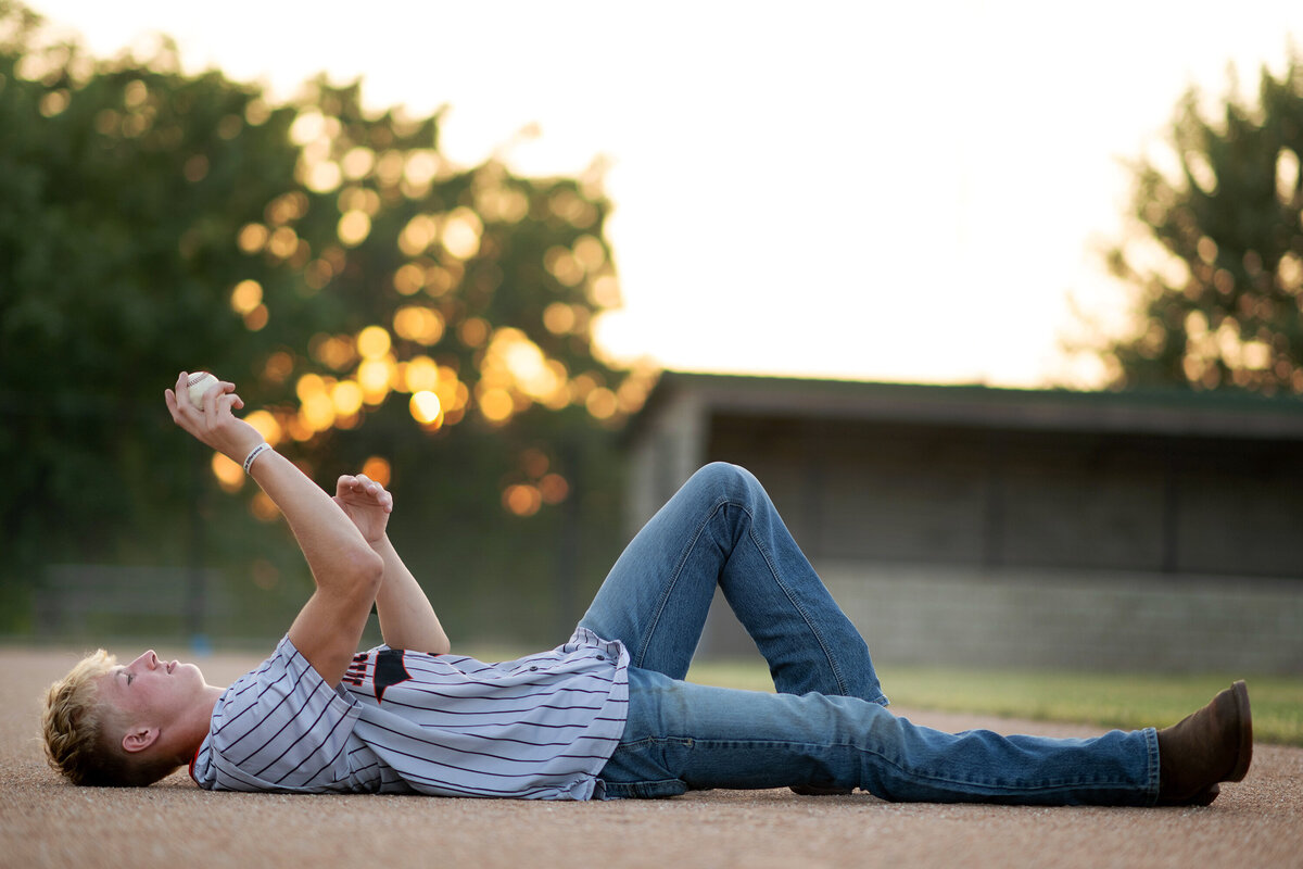A baseball player lays in the dirt tossing a ball up in the air in jeans and jersey shirt at sunset for his Pella Senior Photographer
