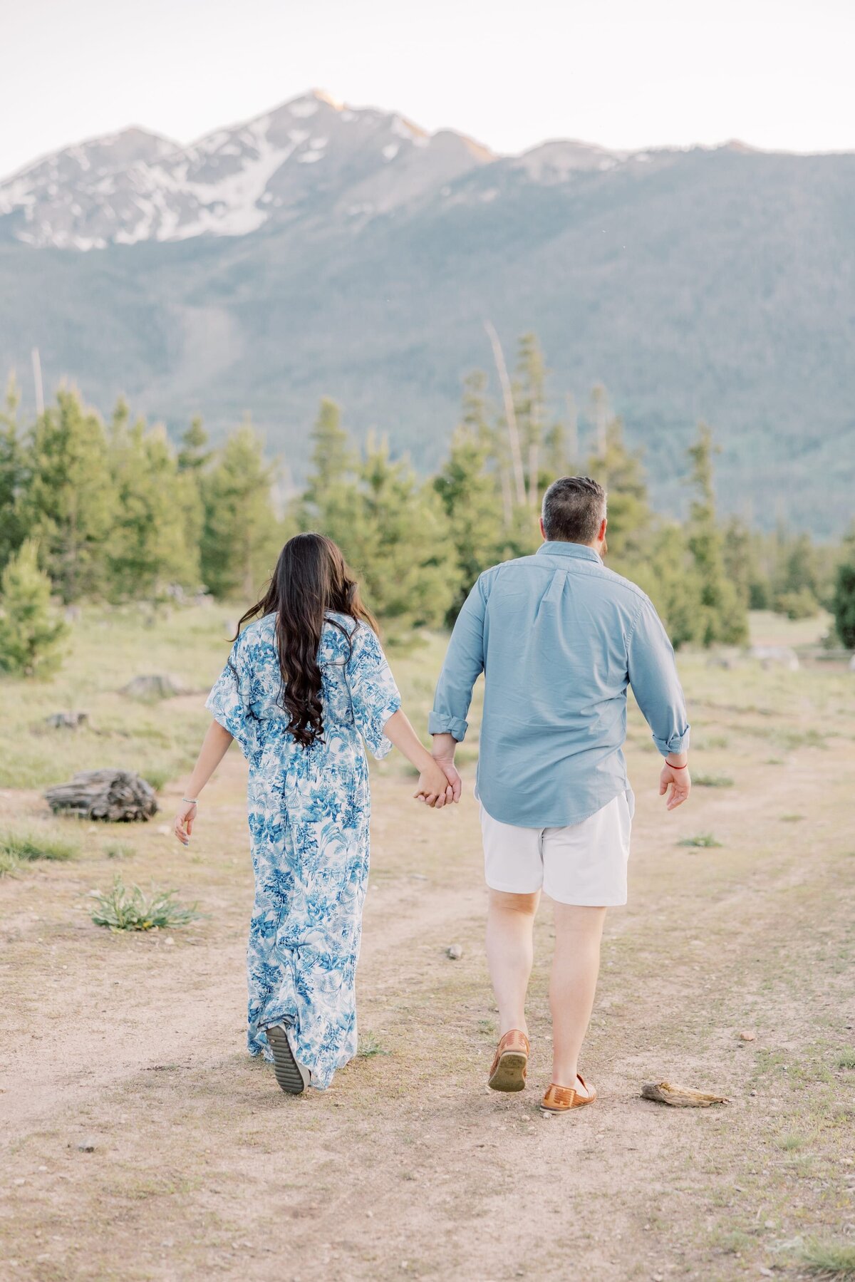 There is a boyfriend and girlfriend walking away from the camera hand in hand. There are pine trees and towering mountain peaks behind them. 