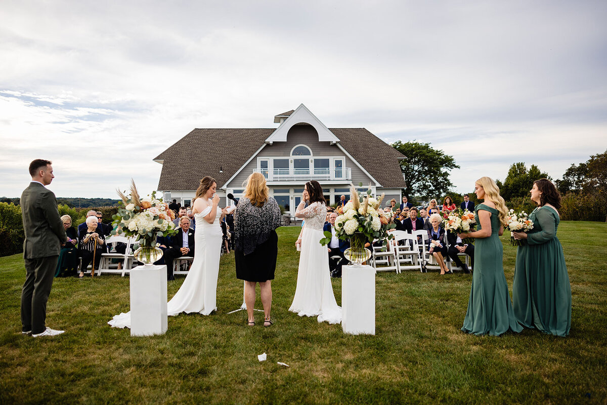 A wedding ceremony in progress with guests seated, overlooking a house. The bride, accompanied by a bridal party in teal dresses