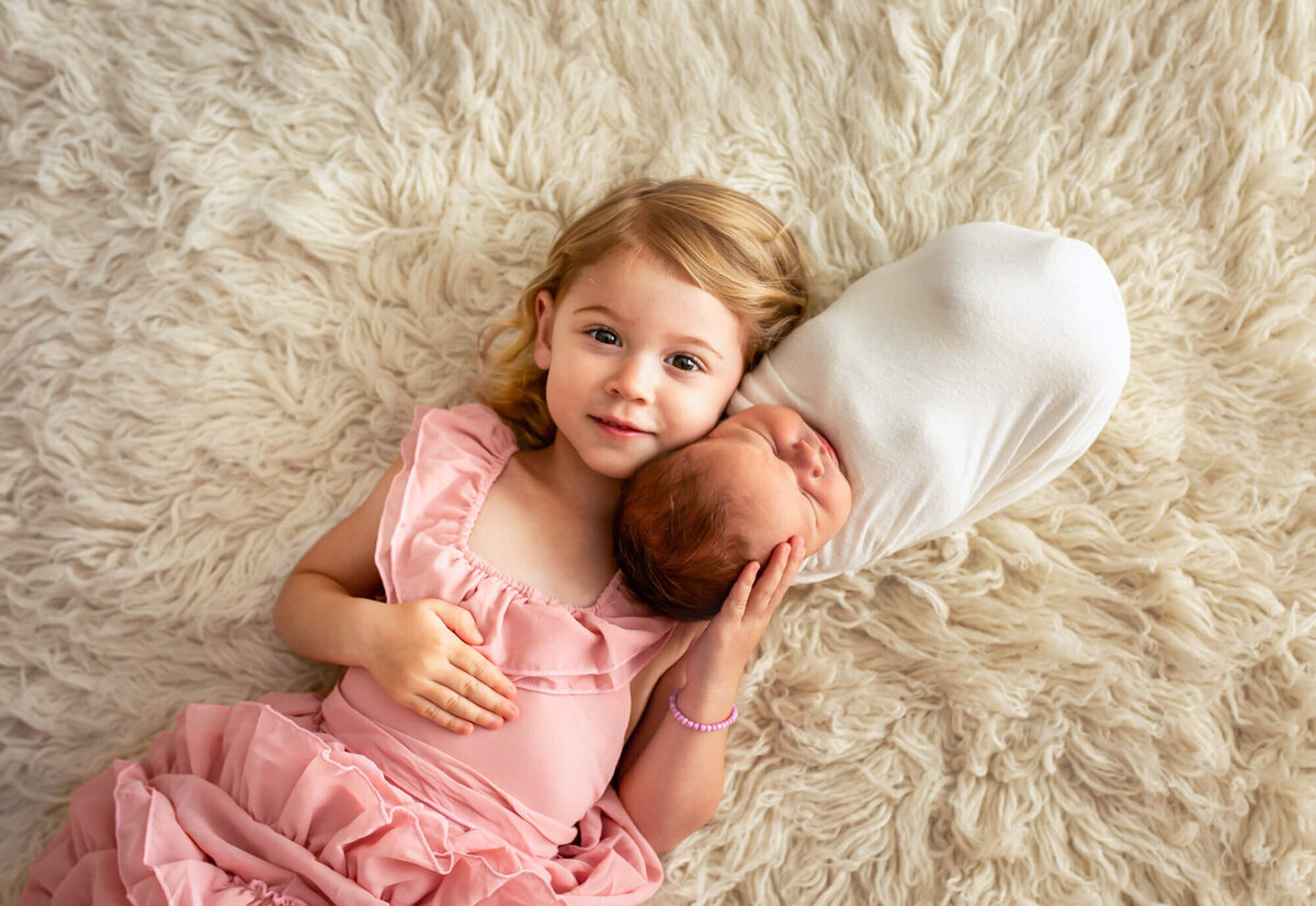 A big sister in a pink dress cuddles her newborn baby brother