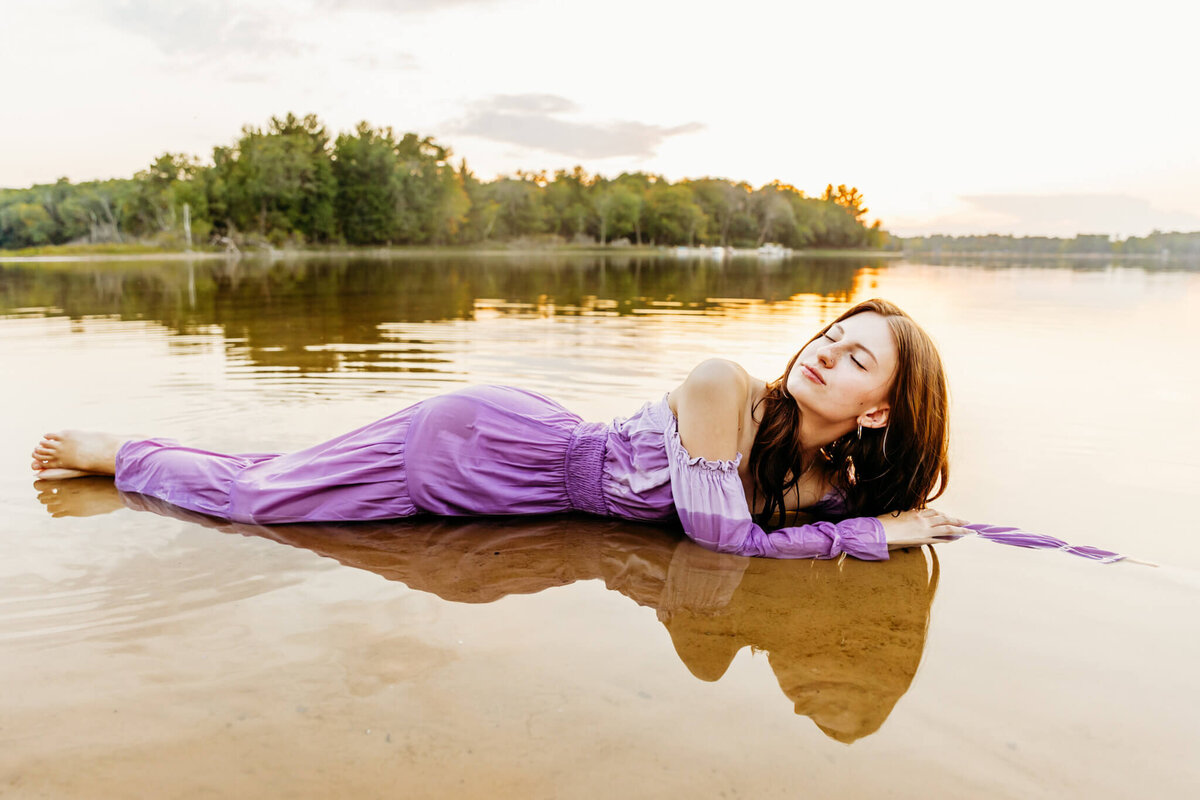 beautiful teen girl laying in a calm lake in her purple dress as she lifts her chin up and takes a deep breath in with her eyes closed captured by Ashley Kalbus
