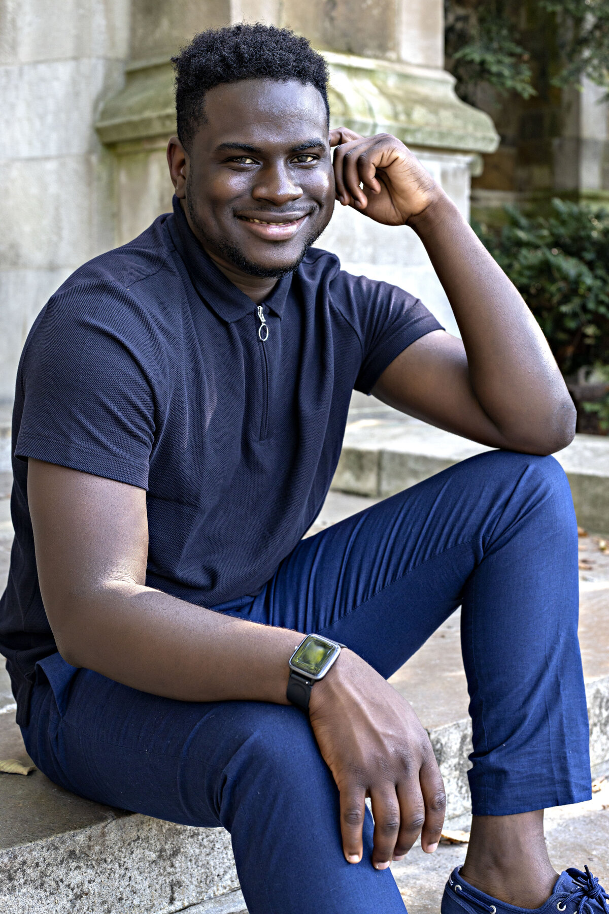 African American male wearing a navy blue zip up top, blue pants, and blue Sperry boat shoes sitting on steps