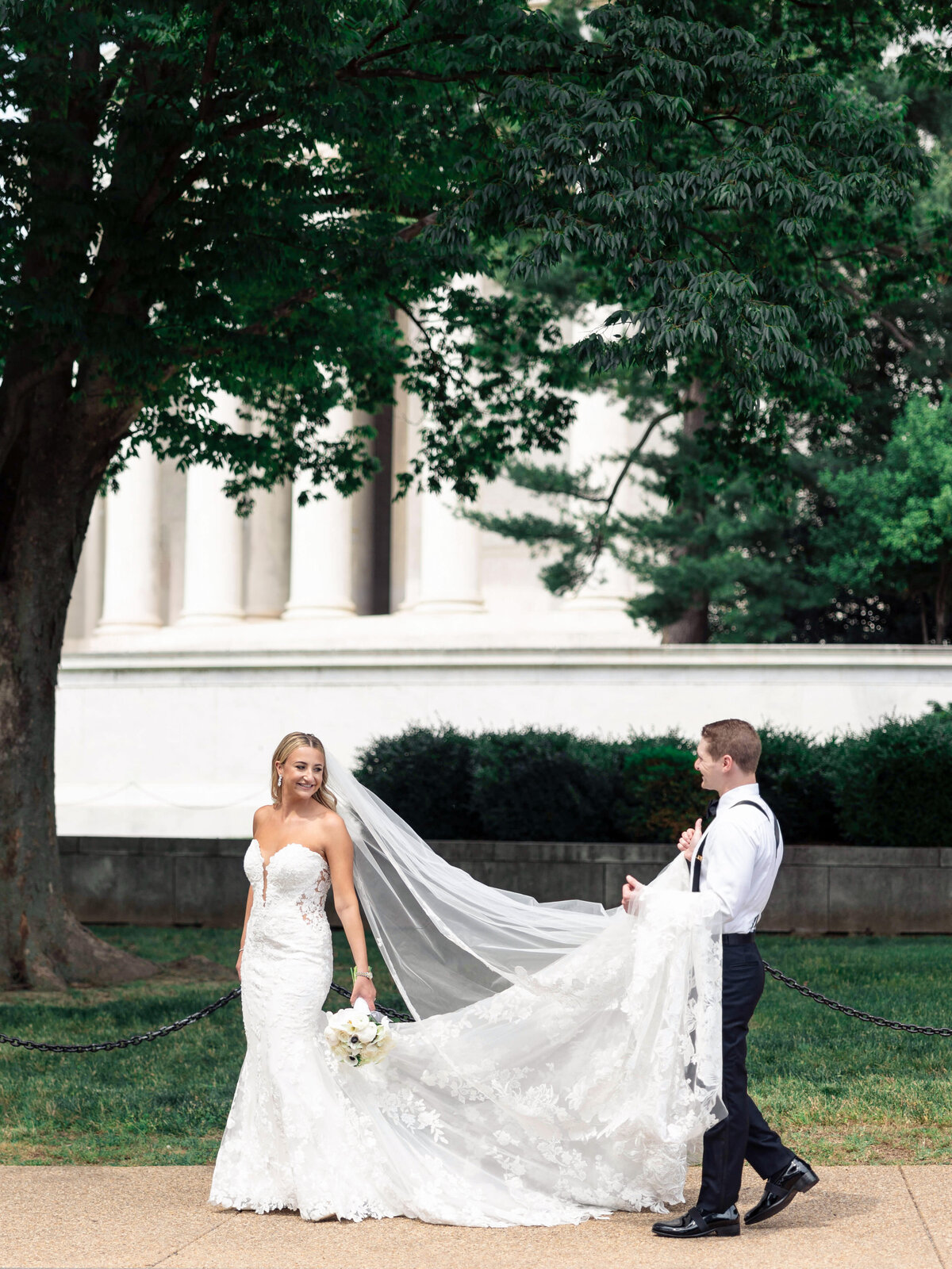A bride in a white lace gown and veil holds a bouquet of flowers while a groom in a white shirt and suspenders helps carry her train. They stand outdoors in front of a large building with columns and greenery.