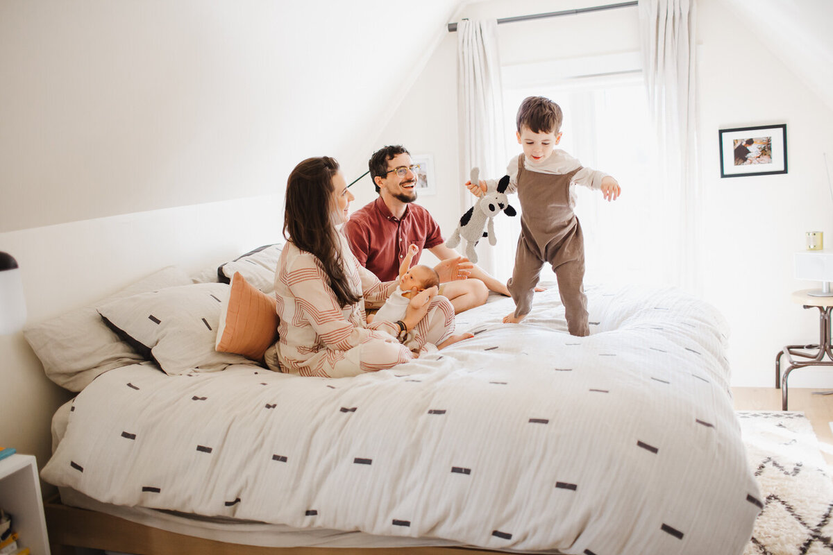 Little boy in brown overalls jumping on bed while parents and newborn baby sister look on smiling