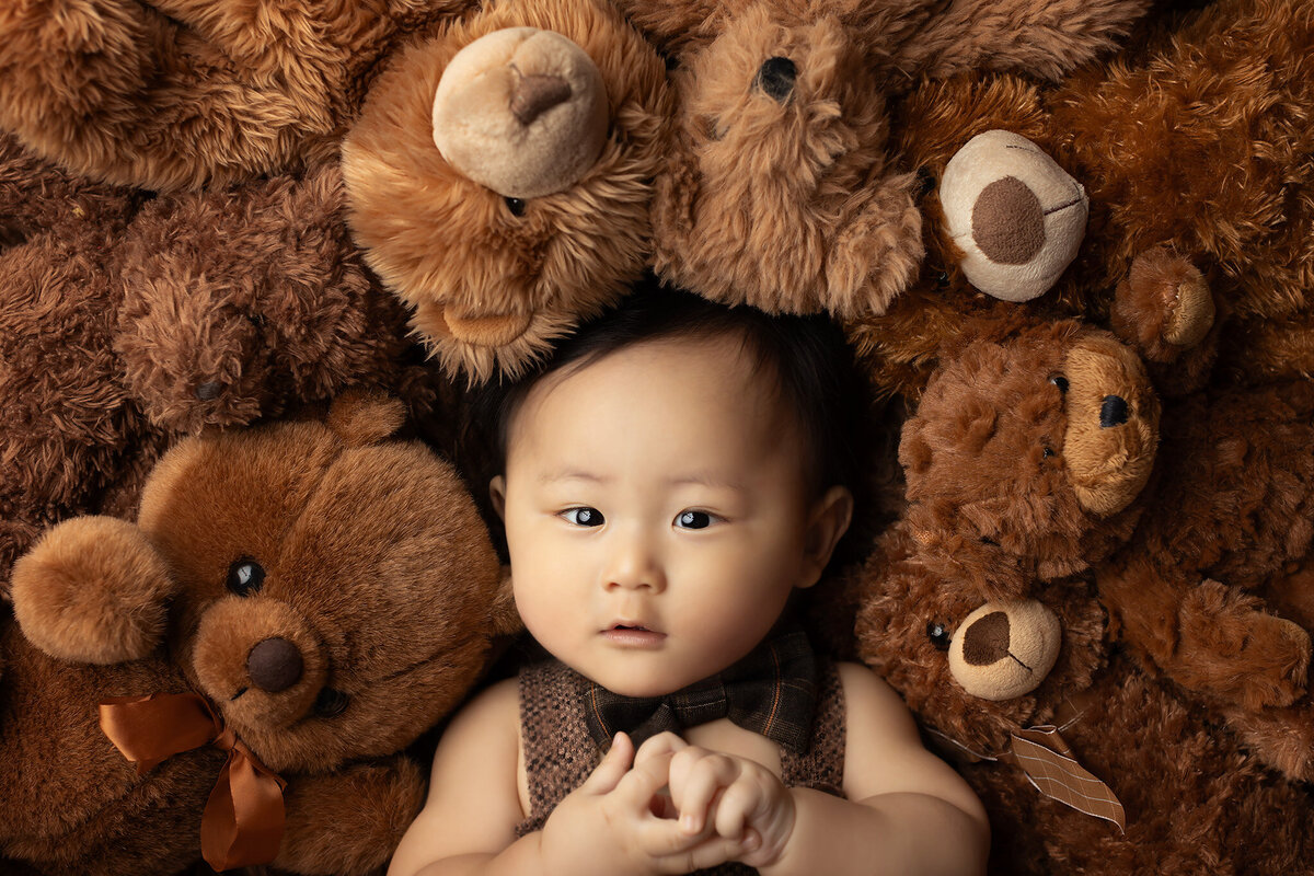 An adorable baby surrounded by a cozy nest of plush teddy bears, looking directly at the camera with wide, curious eyes. This charming milestone portrait by a Dallas baby photographer captures the sweetness of childhood with soft textures and warm tones, perfect for celebrating precious moments.