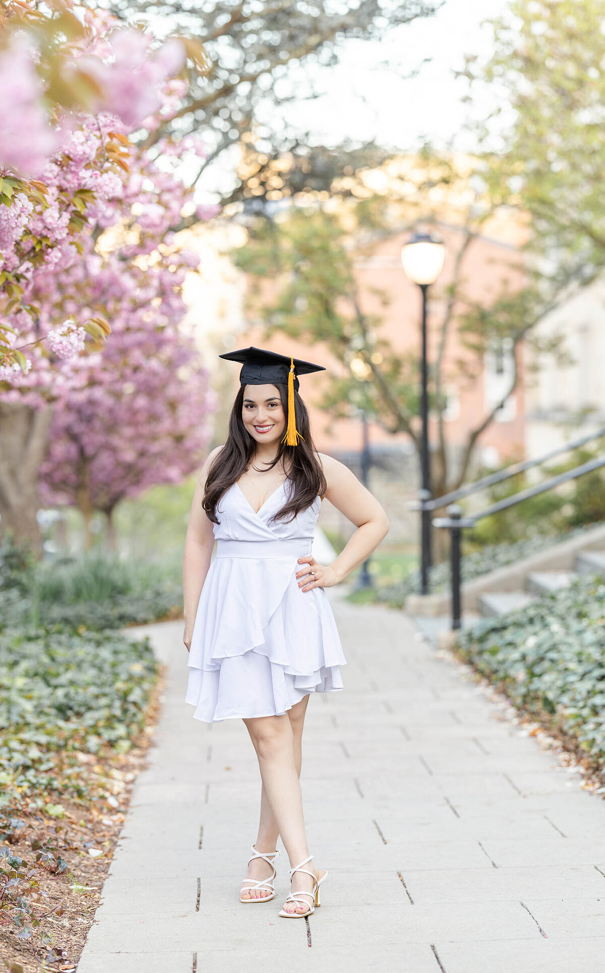 Graduate girl wearing a white stands with one hand on hip smiling at camera during her graduate photo session