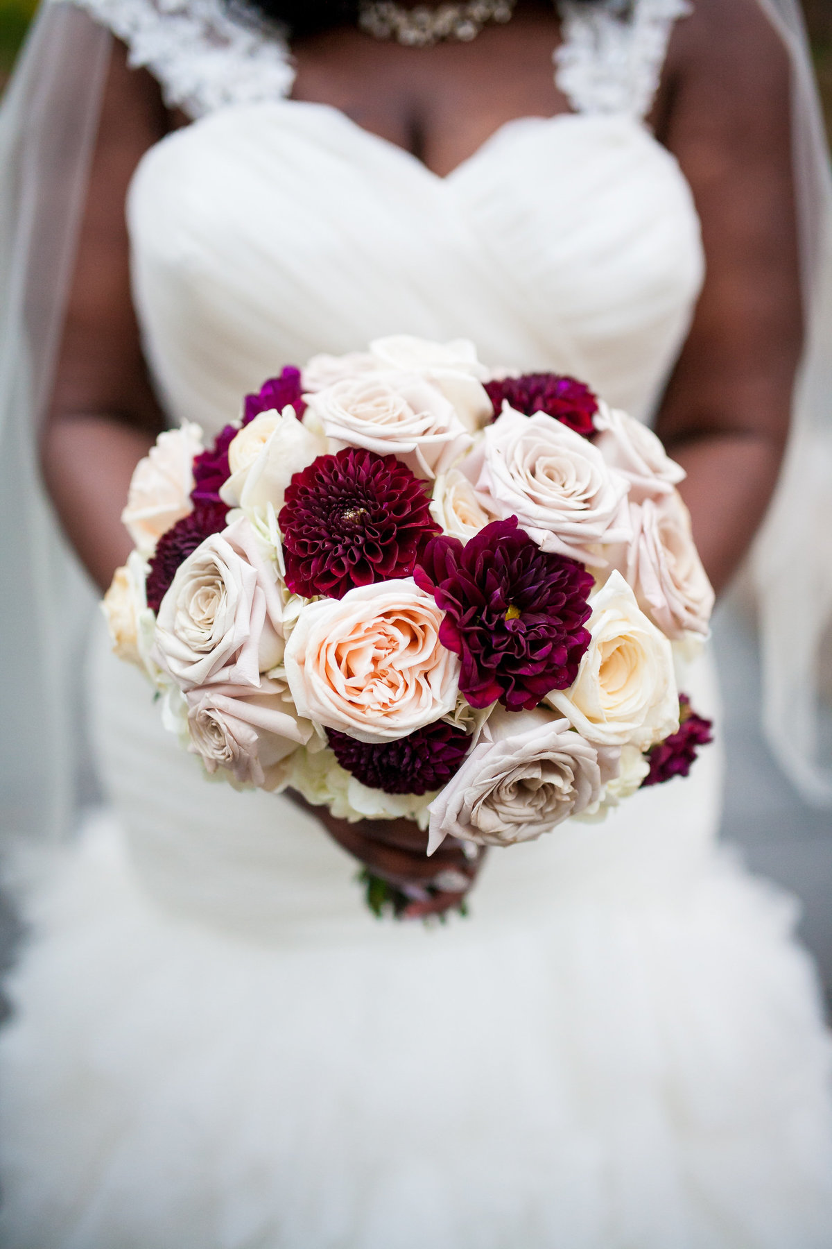 Bride holds her pink, purple, and white bouquet to the camera, her dress becoming the background of this detail photograph.