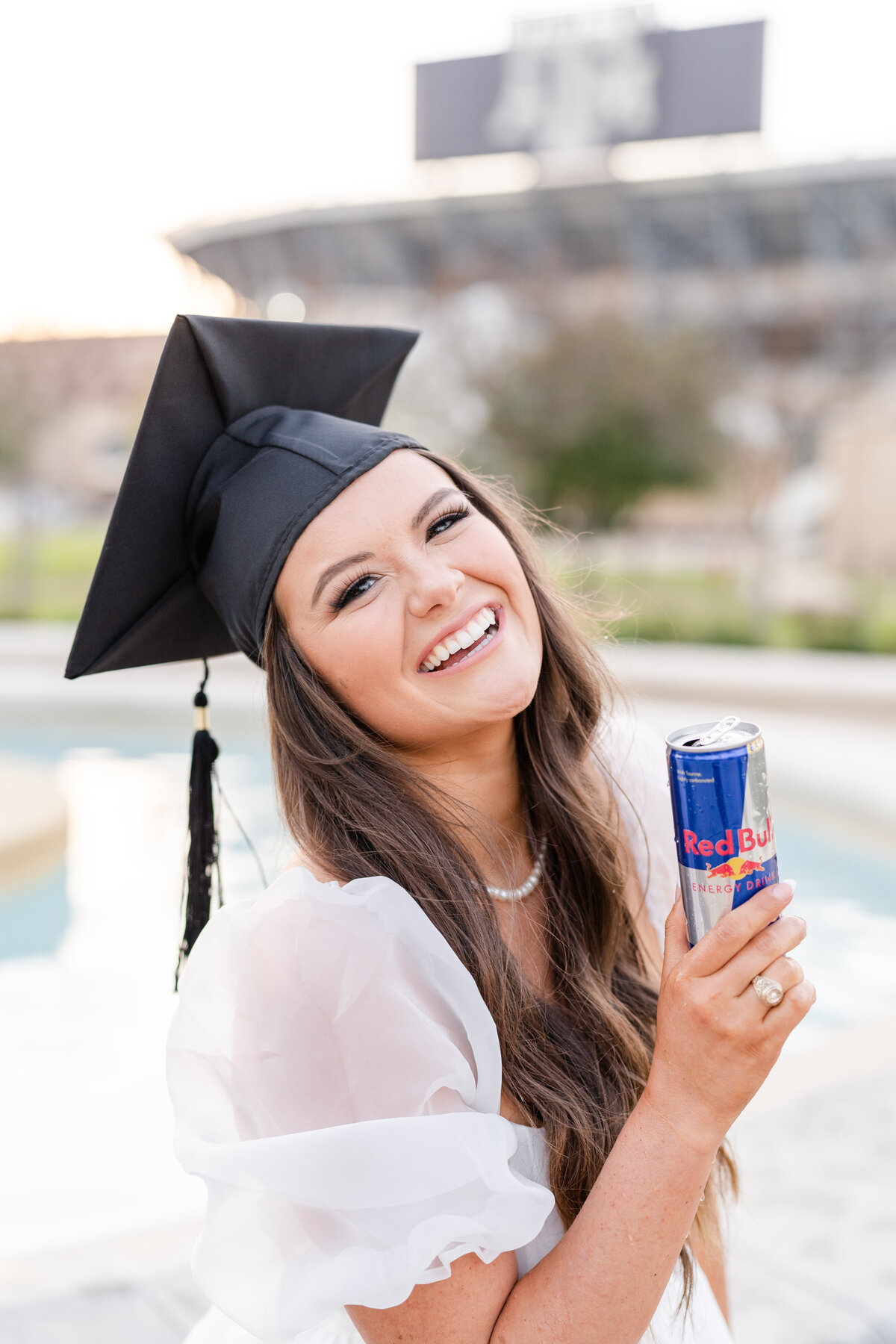 Texas A&M senior girl laughing while holding Red Bull and wearing grad cap and white puffy dress while sitting on edge of fountain in front of Kyle Field