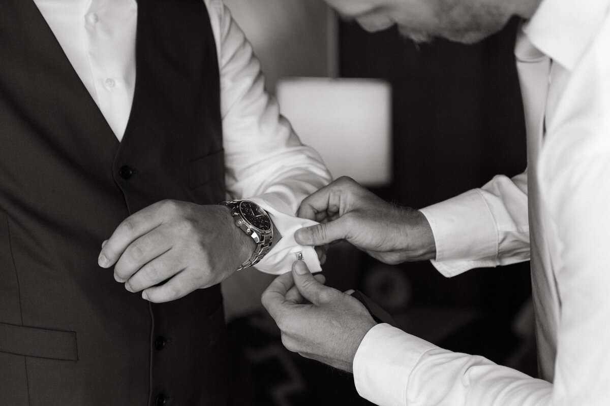 Black and white photo of groomsman helping groom put on cufflinks before his wedding.s