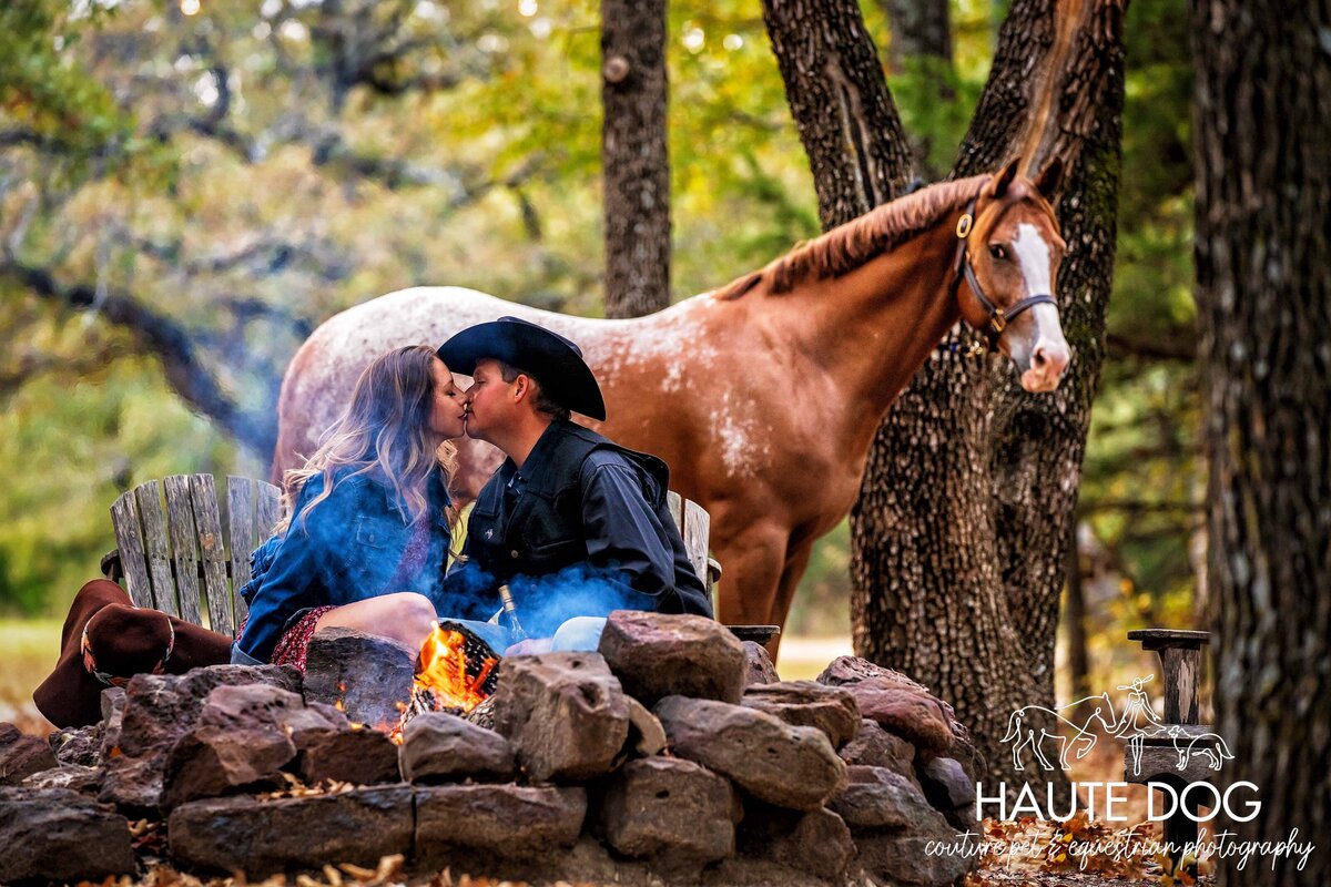 A couple sitting at an outdoor fire pit kiss as their Appaloosa horse stands behind them tied to a tree.