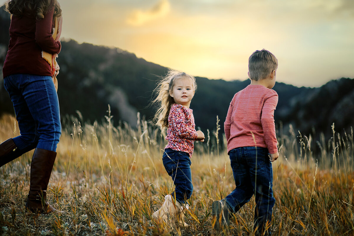 young girl in pink walks next to her brother and mom on a fall day surrounded by tall golden grass and mountains