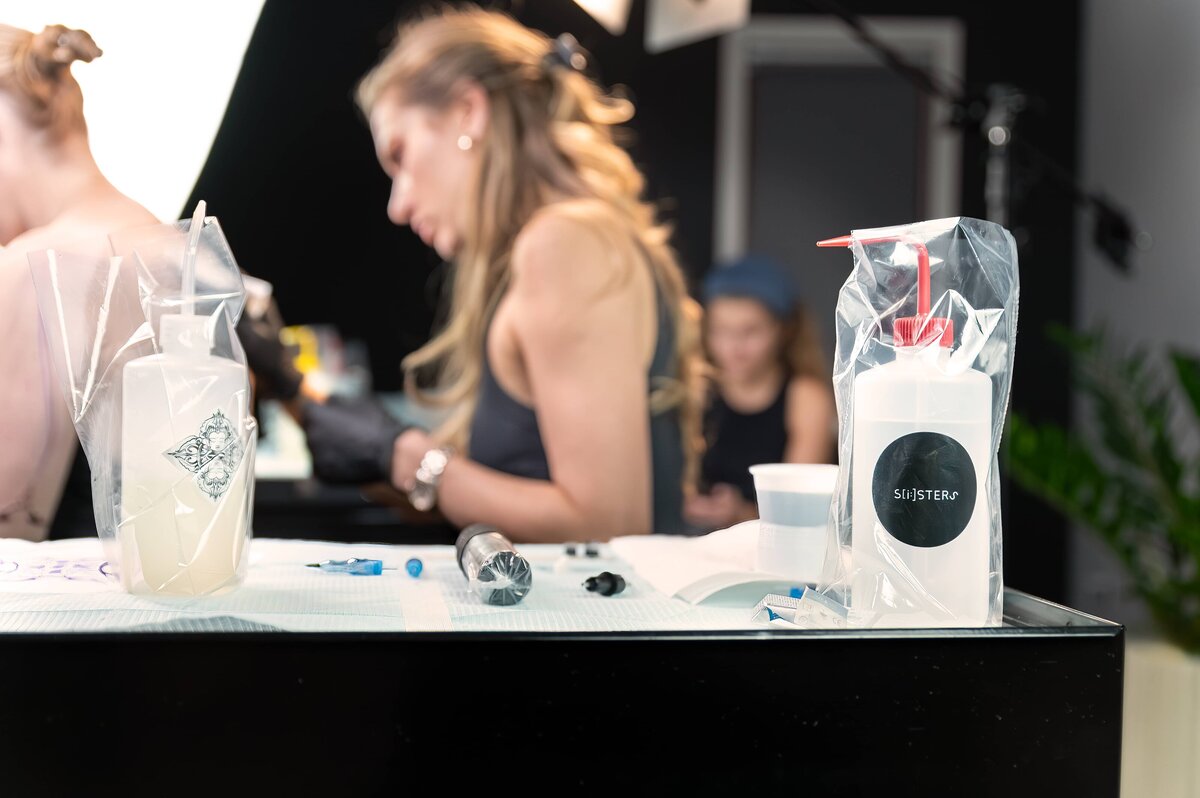 Branded tattoo sanitizing equipment sitting on a cart with tattoo artists tattooing client in the distant background