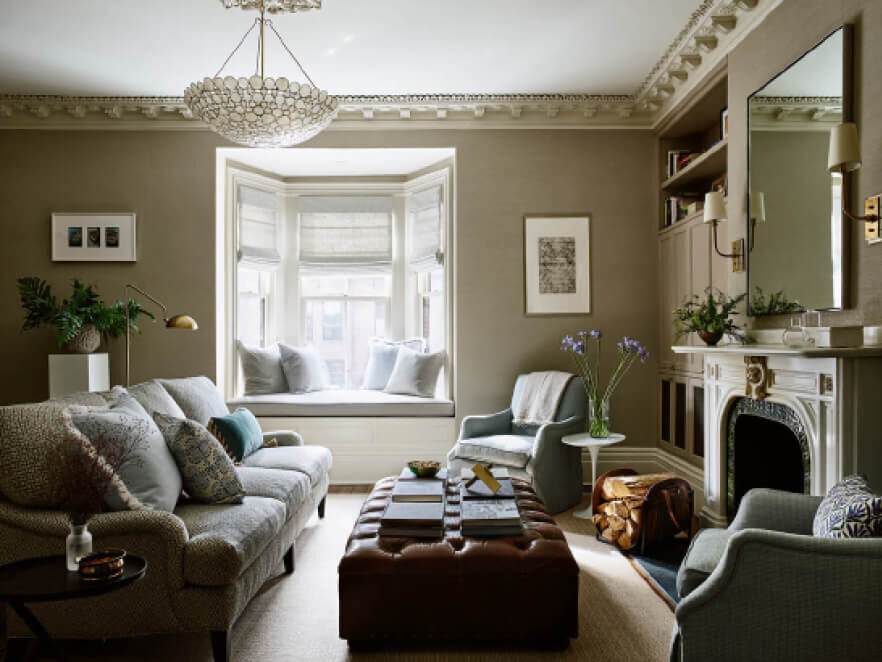 Interior Decorator Boston MA designed a traditional family room in the South End with grasscloth wallpaper, an Oly chandelier, a leather tufted ottoman, and a textured fabric sofa.