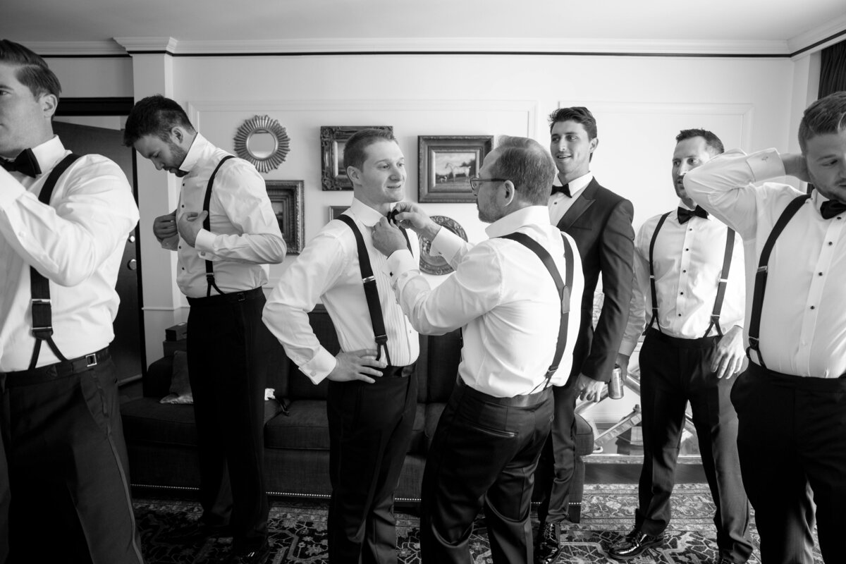 A group of groomsmen in a room getting ready for an event. They are wearing formal attire with suspenders and bow ties. One older man is adjusting a younger mans tie. The atmosphere is lively and focused. Black and white image.
