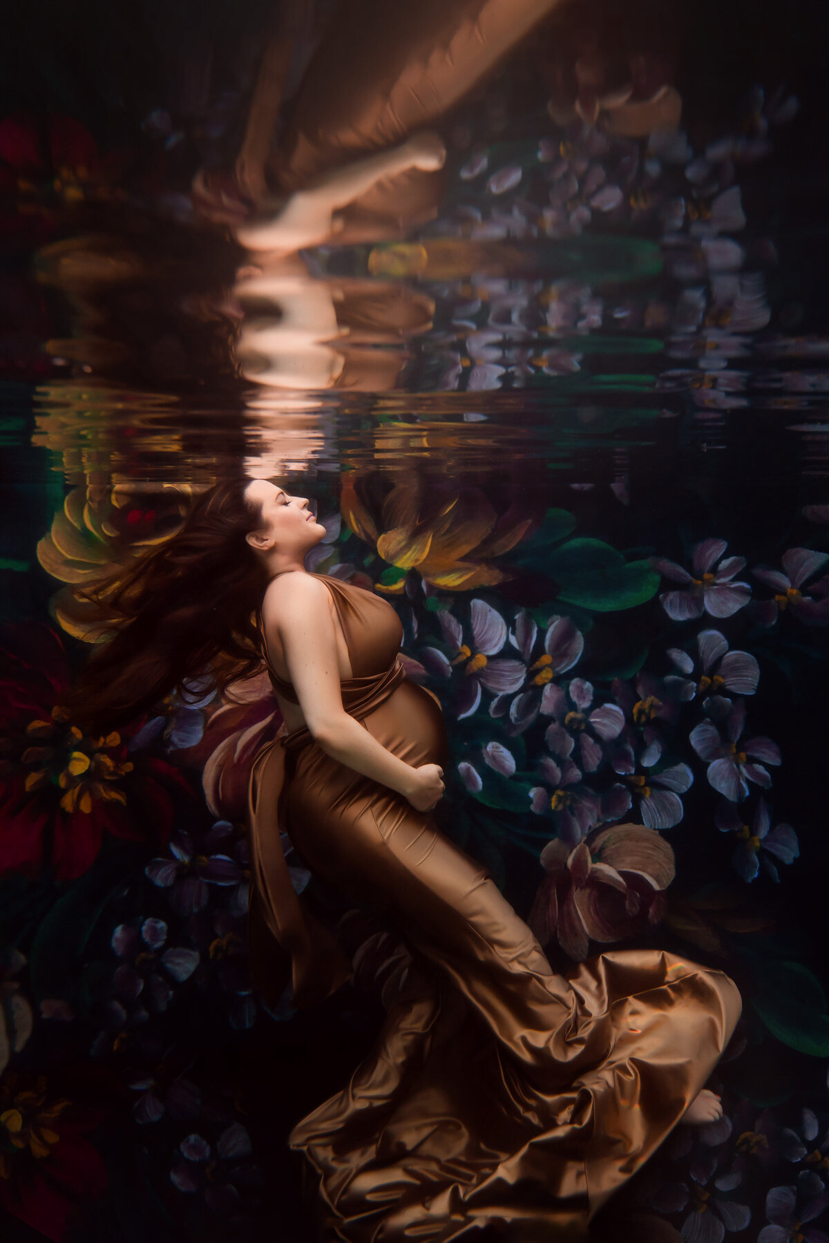An enchanting underwater maternity photo featuring a woman in a luxurious golden gown surrounded by vibrant floral art. The elegant pose and dreamy reflections highlight this captivating Dallas underwater maternity photographer's artistry.