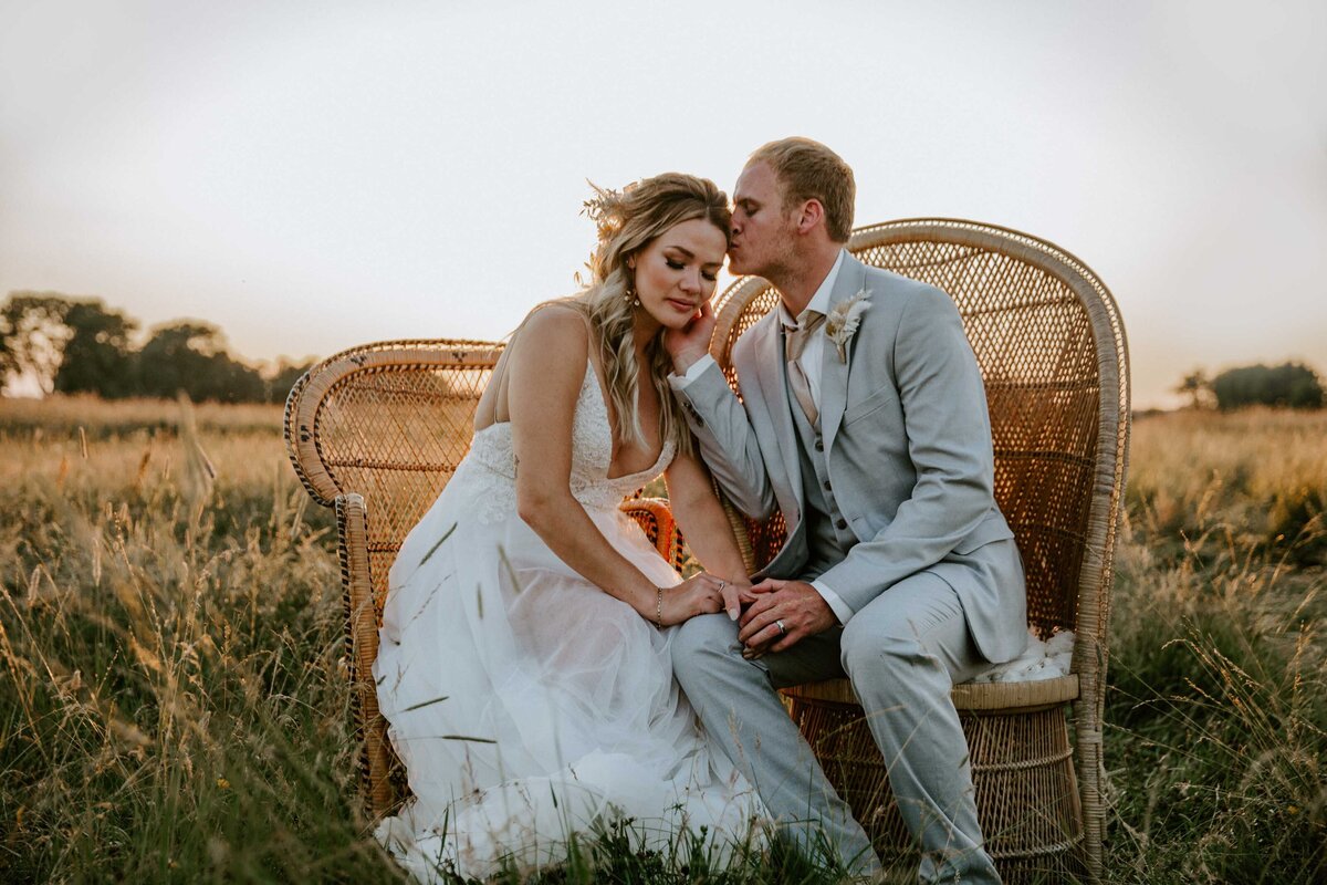 Bride and groom are sitting in vintage fan chairs in Exeter, Ontario field for intimate wedding photo. The bride is leaning into her groom. The groom is touching her cheek and kissing her forehead.