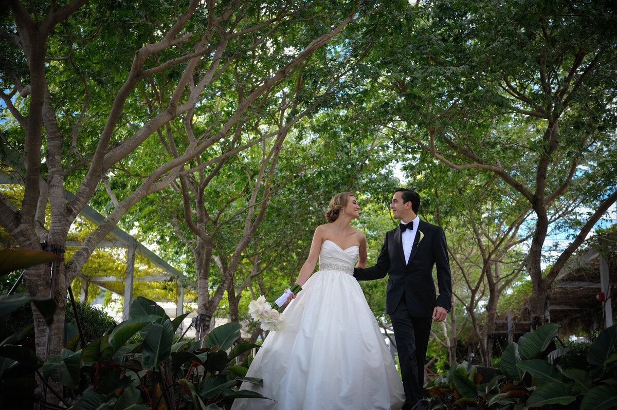 Bride and groom walking together under lush trees in a garden setting, Miami, Florida.