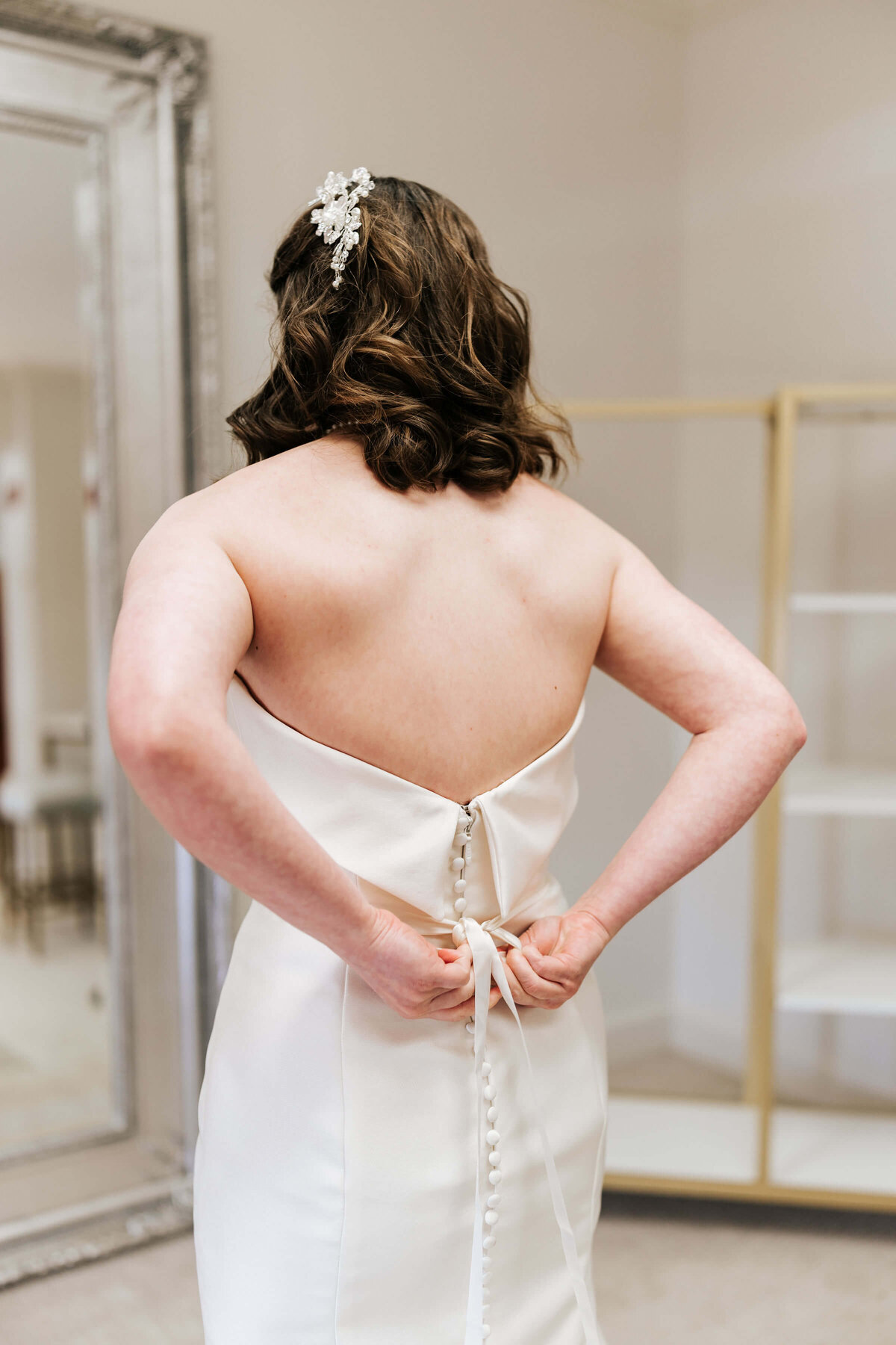 A woman in a strapless white dress ties the back of her gown. Her hair is styled in loose waves with a jeweled hairpiece. She stands in a fitting room with a large mirror and empty shelves in the background.