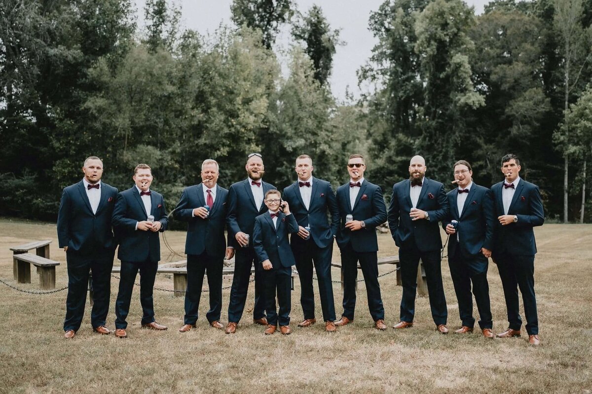 A group of groomsmen in navy suits and brown shoes lined up in a forested area, smiling confidently towards the camera.