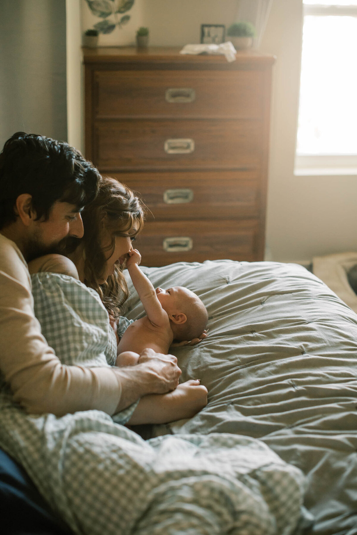 Mom and dad lying on bed with newborn sun with sun through window Newborn Portrait Photography