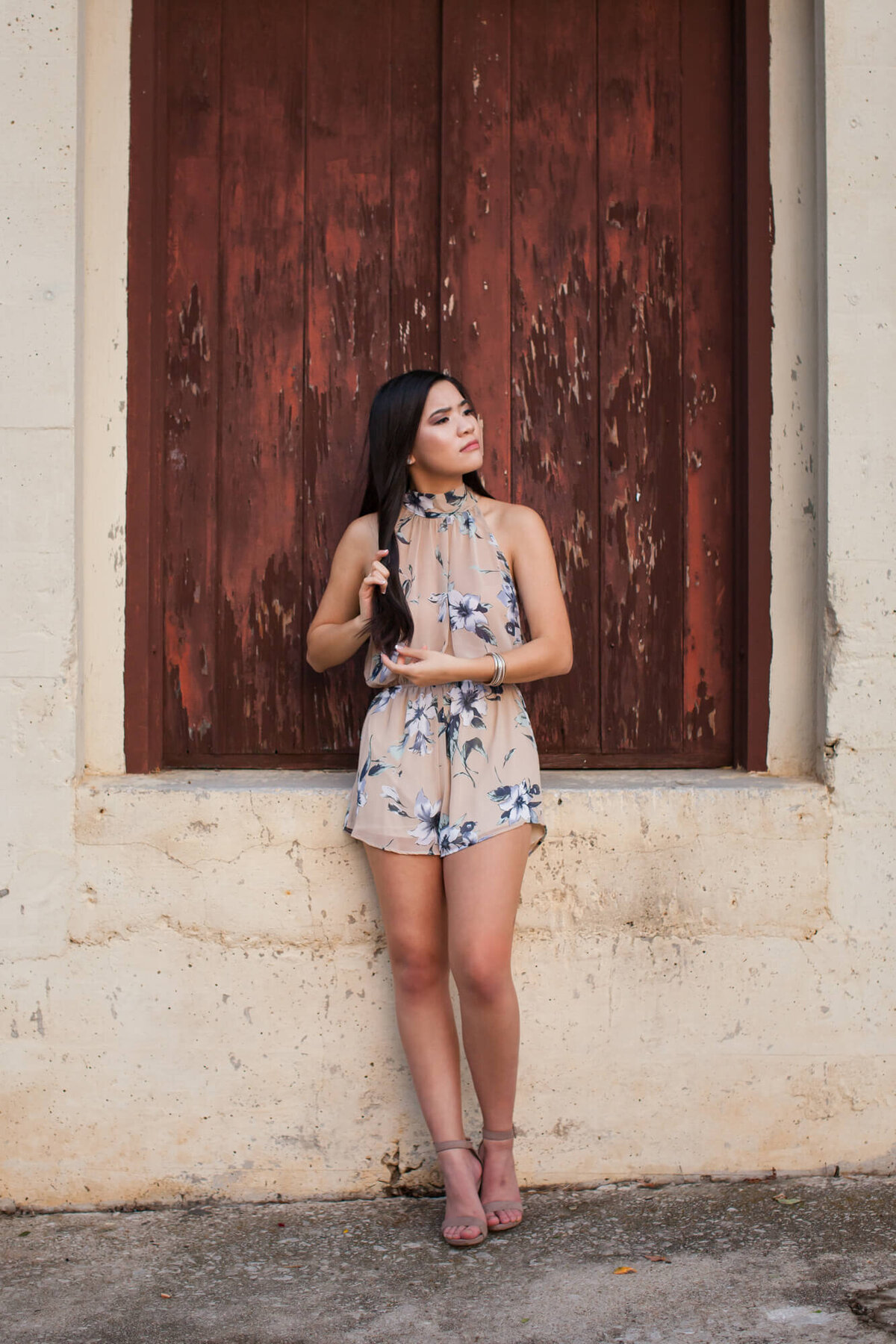 A lovely black haired senior girl poses casually in front of a rusty colored wooden shuttered window. Captured by Springfield, MO senior photographer Dynae Levingston.