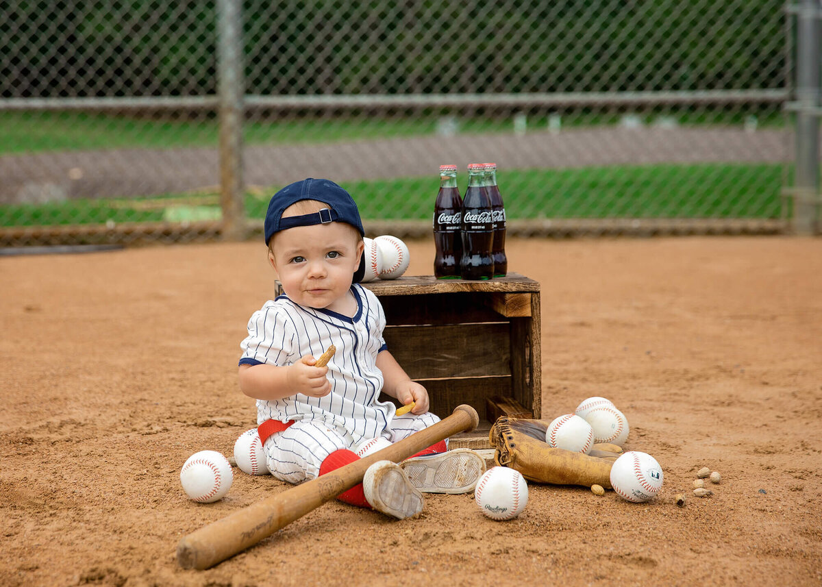 1 year old boy sitting on a ball diamond with coke, peanuts balls, bat and glove around him. He's wearing a baseball outfit with his hat turned backwards.