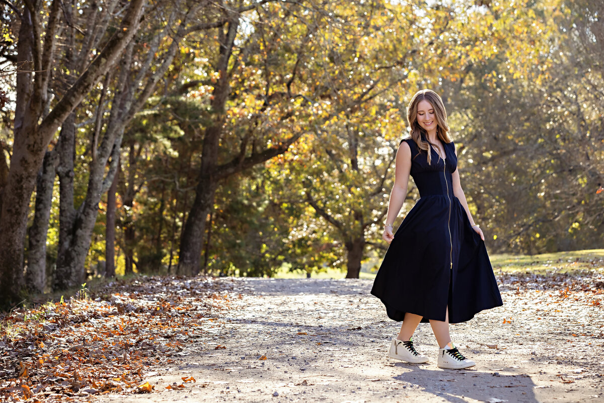 High school senior in black dress walking dreamily along gravel path lined with yellow trees