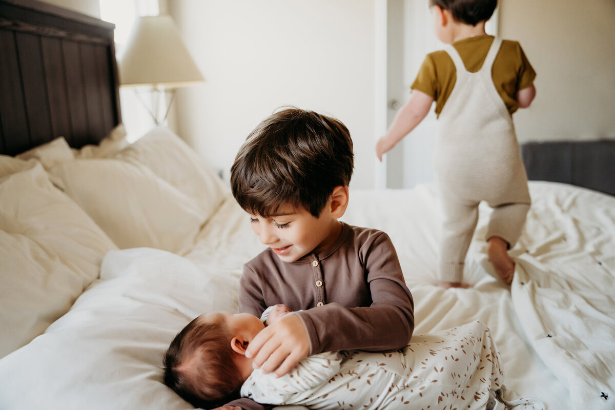 Newborn Photographer, older brother admires new baby brother on the bed, another sibling plays on the bed