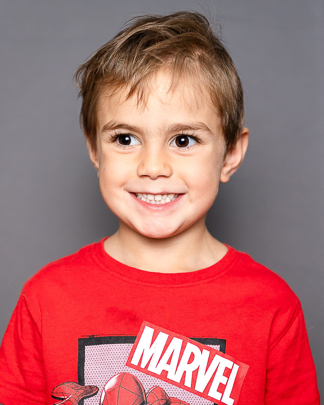 Young child smiling, wearing a red t  shirt, standing against a gray background.