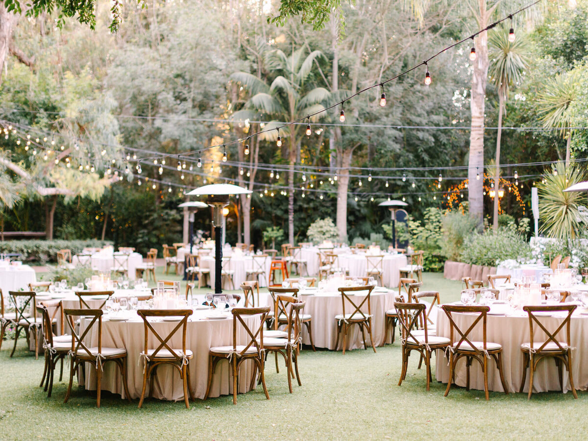 An elegantly set outdoor dining area with round tables, string lights, and lush greenery, perfect for a wedding.