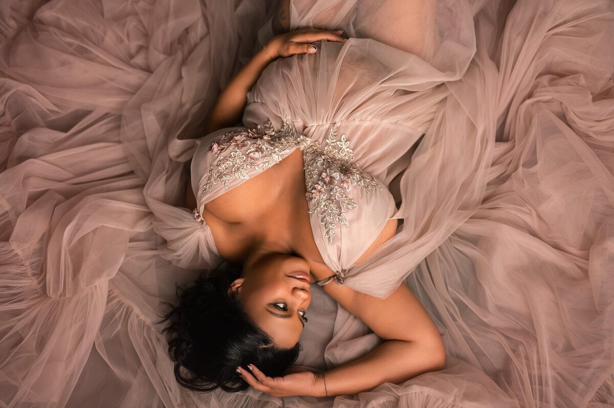 Pregnant woman lying gracefully on a bed of soft, flowing tulle in a blush embroidered gown, captured in a warm and elegant studio setting in Waukesha, Wisconsin.
