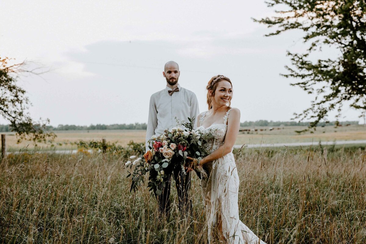 Groom stands in the distance behind his bride for formal wedding photo. Bride is in the foreground holding her bouquet looking off in the distance at a rustic barn wedding in Exeter, Ontario.