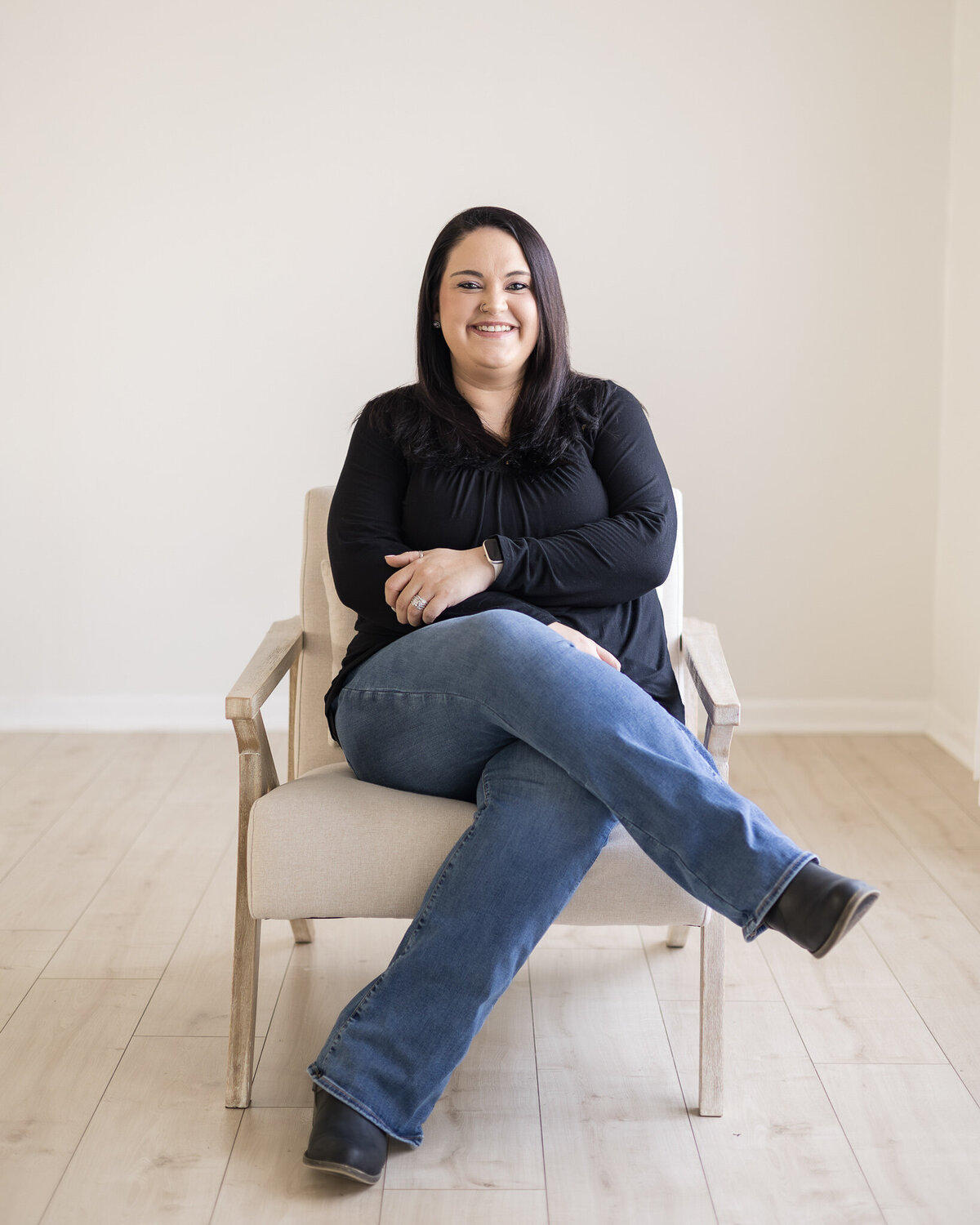 Woman sitting in a chair with legs crossed, has black hair, wearing a black shirt, blue jeans, and black shoes, smiling at the camera