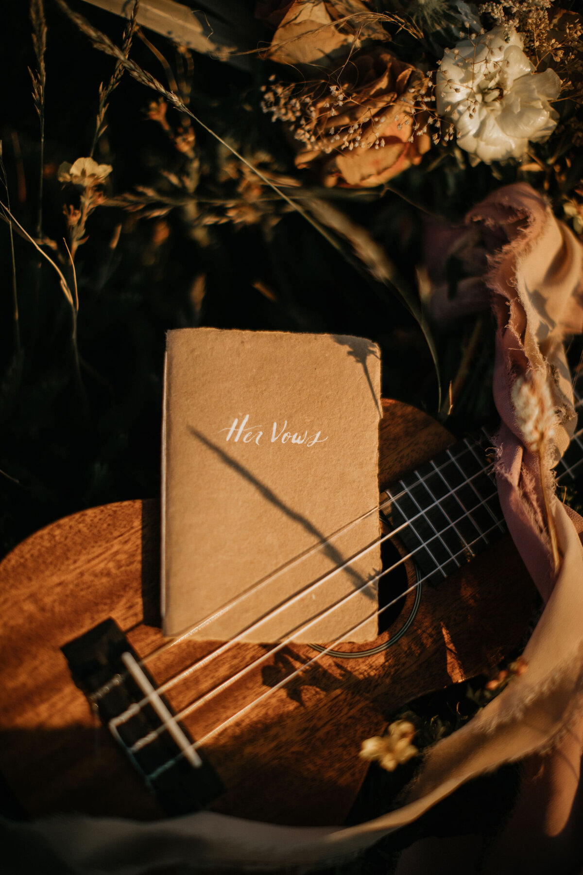 Tan wedding stationery with a white cursive font that reads ‘her vows’ set atop a ukulele with flowers