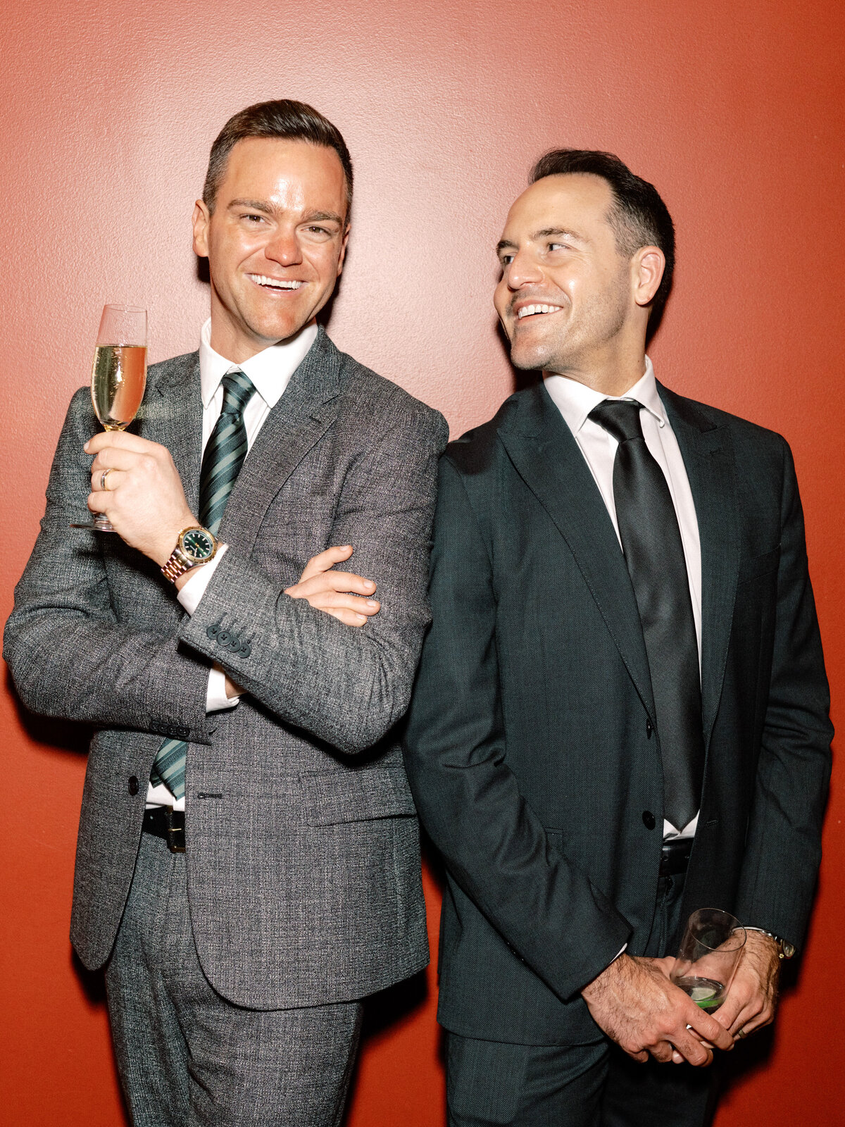 Two sharply dressed men enjoy a moment together, sharing smiles and drinks at a sophisticated event, embodying elegance and good company.