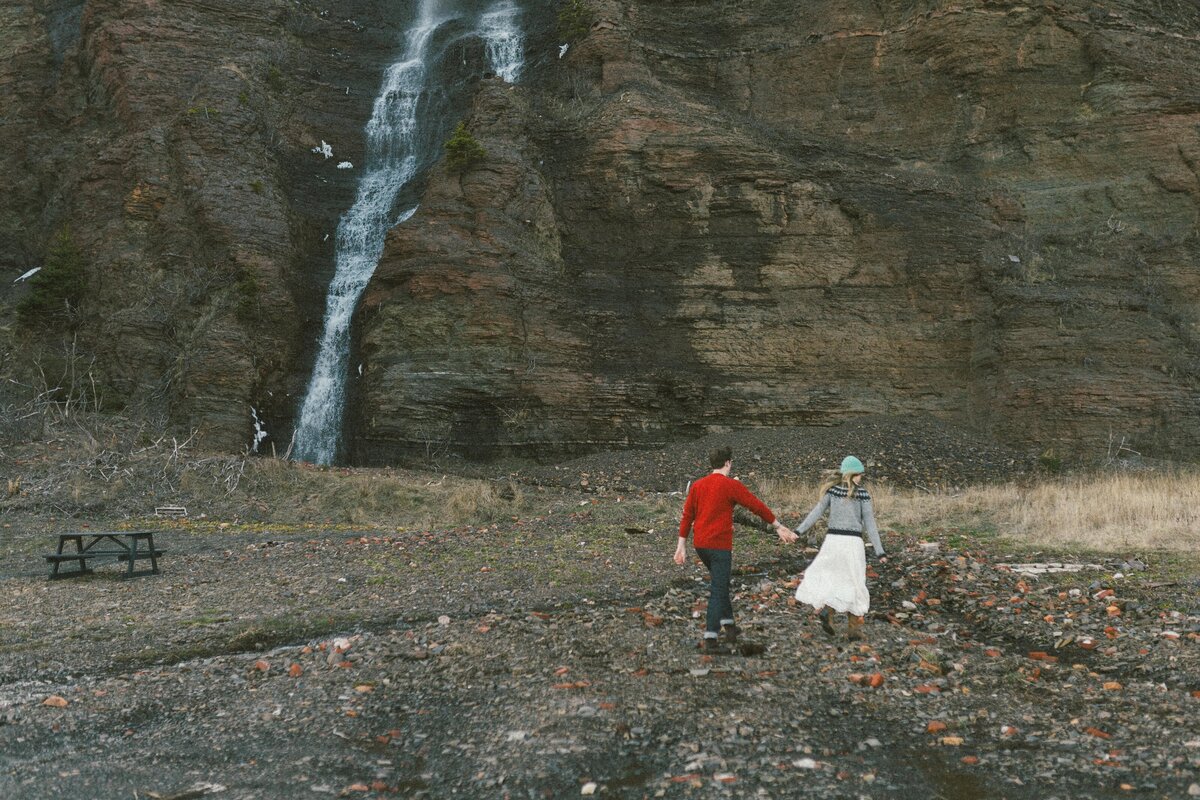 bride and groom hold hands and walk through rocky landscape at the base of a waterfall