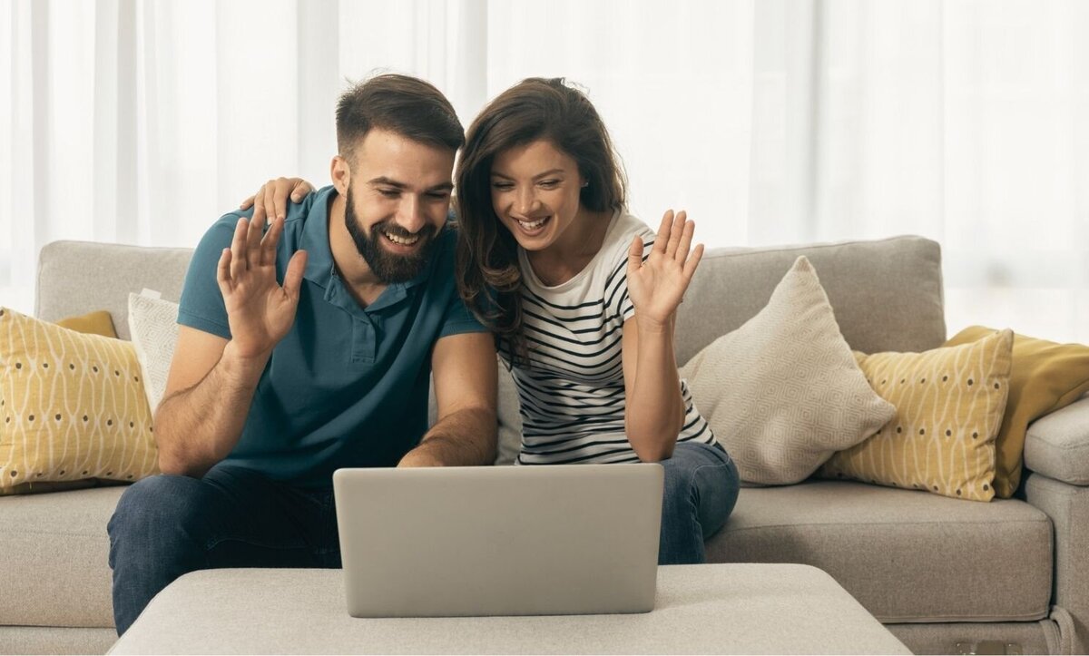 A couple sitting on a couch smile at their laptop as they wave. This could represent a couple meeting with an online therapist in Florida over a teletherapy platform. Contact an online therapist for support with online relationship therapy and other services.